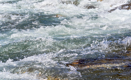 Rapid spring river flowing over rocks on sunny day, forming white water waves, closeup detail -...