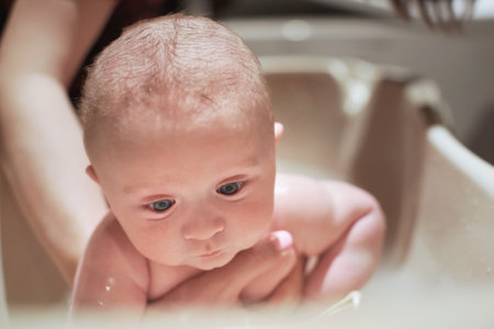 Infant baby boy washed in small bath tub, detail on his face as mother holds him under belly