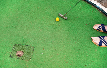 Young man playing minigolf, closeup view from above on his feet in sandals, yellow ball, metal club and worn green floor near holeの写真素材