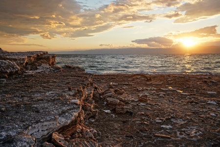Sun shines through dramatic sunset clouds to waves at Jordanian side of Dead Sea, branches, flotsam, debris and rubbish washed on shoreの写真素材
