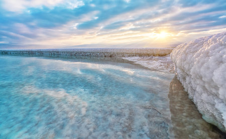 Sand completely covered with crystalline salt looks like ice or snow on shore of Dead Sea, turquoise blue water near, sky colored with morning sun distance - typical scenery at Ein Bokek beach, Israelの写真素材