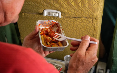 Passenger eating airplane food, pasta and sauce from aluminium container, wit plastic fork, detail...