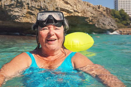 Elderly senior woman smiling in calm calm sea on sunny day, fogged diving mask on head, rocky cliff...