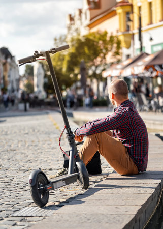 Young man in casual trousers sitting on sidewalk next to his parked electric scooter, blurred city backgroundの写真素材