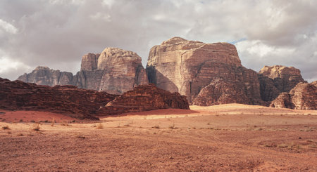 Rocky massifs on red sand desert, few dry grass cluster ground, cloudy sky in background, typical scenery in Wadi Rum, Jordanの写真素材
