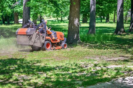 Man mows the grass under trees in park by riding mowing machine in sunny day の写真素材
