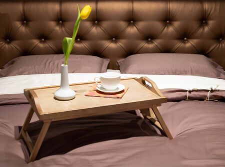 Wooden tray of coffee and candles with flowers on bed. Brown bedding sheets with striped blanket and...
