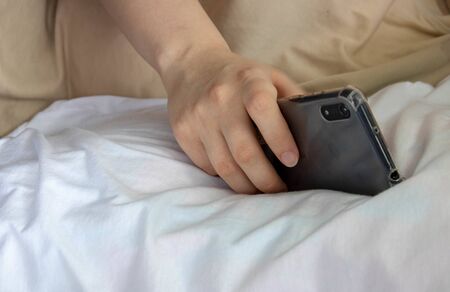 Woman hand under blanket being woken by mobile phone in bedroom.の写真素材
