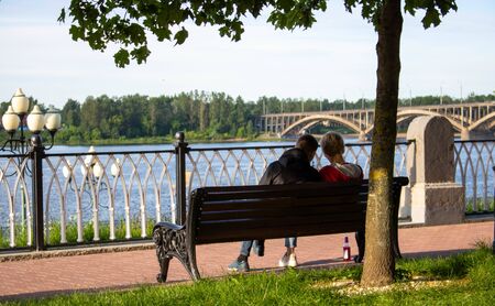 A young couple is sitting on a wooden bench under a tree near the river.の写真素材