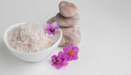 Pink coarse salt in a white bowl stands on a white background next to pink stones and pink violet flowersの写真素材