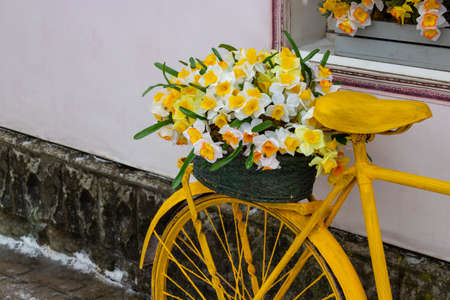 A yellow bicycle wheel with a basket of flowers stands against the wall, in a vintage style.の写真素材
