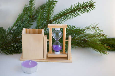 On a white background, a transparent hourglass with flowing lilac sand, a candle and fir branches....