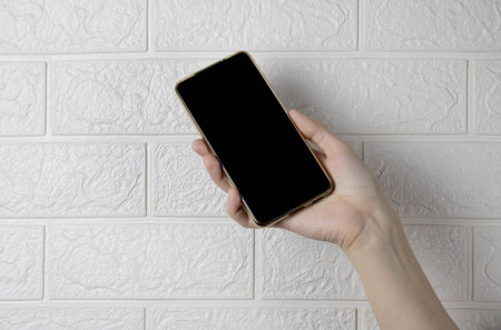 A hand holding a new smartphone on a white brick wall background