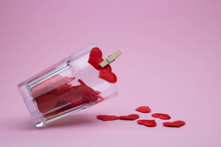 An overturned glass glass on a pink background is filled with red heartsの写真素材