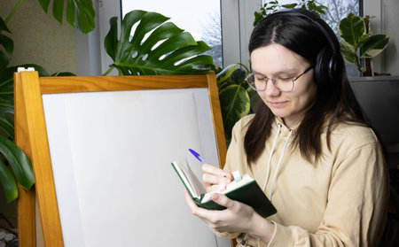 A student girl in glasses and headphones writes in a notebook against a background of home flowers, windows, next to an easelの写真素材