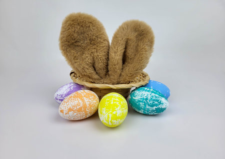 Brown rabbit ears stick out of a straw hat and colorful Easter eggs are lying next to it. The...
