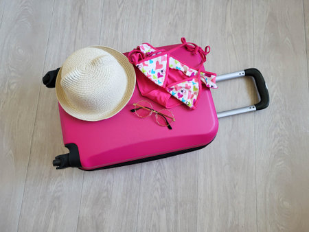 On the pink children's suitcase are a children's swimsuit, hat and sunglasses. A suitcase on wheels with a handle is lying on the floor.の写真素材