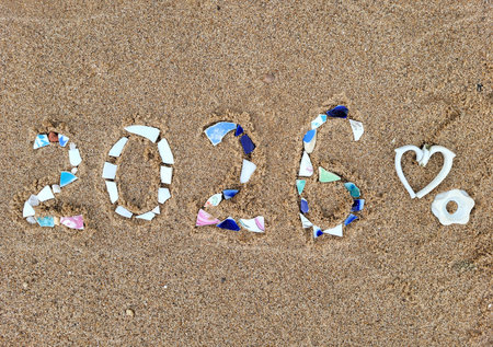 The numbers 2026 are written on a sandy beach with colorful pieces of dishes. The concept of New...