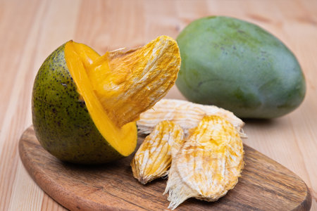 A ripe large yellow mango fruit is cut in half, a large mango stone is visible, lying on a wooden tray. Nearby on a wooden table there are large mango seeds and a whole mango fruitの写真素材