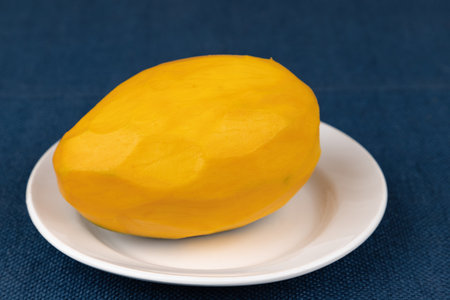 Peeled whole ripe mango fruit of yellow color lies on a white plate on the table.の写真素材