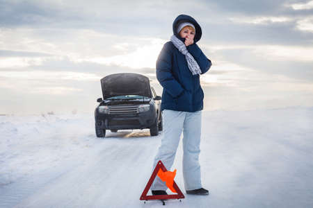 The car broke down on the road. The woman warms her cold hands. She's cold. Winter. Emergency stop sign. Around a snowy field.の写真素材