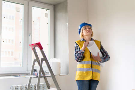 A female Builder looks at the drawings of the object. A middle-aged woman in a helmet makes repairs in the apartment.の写真素材