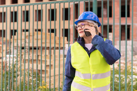 Construction worker woman on construction site in green vest and helmet with phone. A middle-aged...