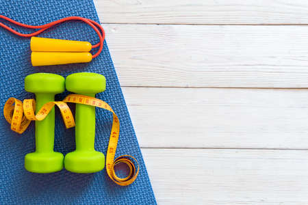 Green dumbbells with a jump rope on a mat on a white wooden background. The concept of fitness and a...