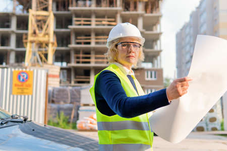 A female construction worker in a vest and helmet on the background of a new building, She looks at...