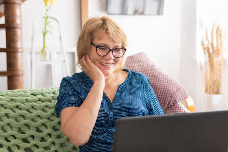A woman with a laptop is sitting on the sofa. She watches the news and talks to her friends