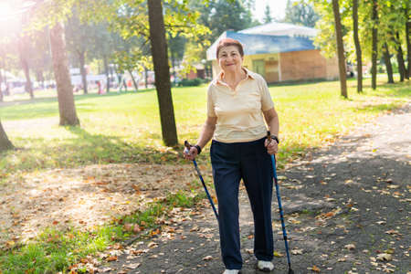 A woman in the Park walks nordic with sticks on a Sunny summer day. Senior woman with a good mood is engaged in morning work-out.の写真素材