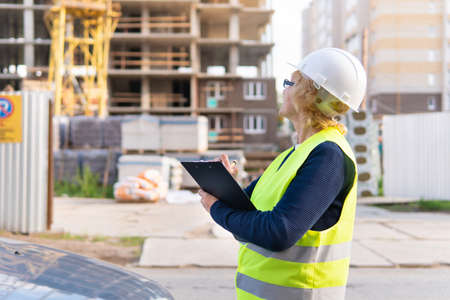 A female construction worker in a vest and helmet writes in a notebook. She's inspecting the...