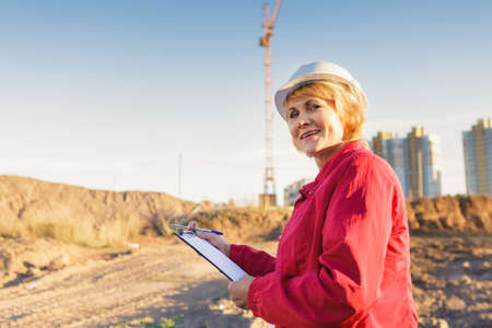 A female construction worker is on a construction site. A middle-aged woman in a white helmet with a tablet makes an inspection of the territory.の写真素材