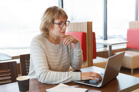 A woman in a cafe is working on a laptop. She smiles. A middle-aged woman, an adult in an office. She watches social media.の写真素材