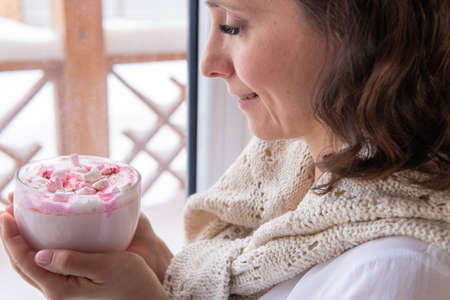 A pretty woman drinks a drink with marshmallows by the window.