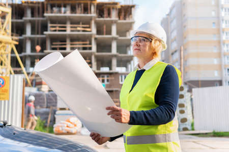 A woman at a construction site in a blue suit, vest and helmet. A middle-aged woman with a project and drawings inspects the construction.の写真素材