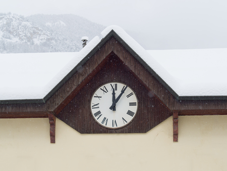 wall clock with Roman numerals, on the facade of an old house in the mountains, marking  five minutes after noon while snowingの写真素材