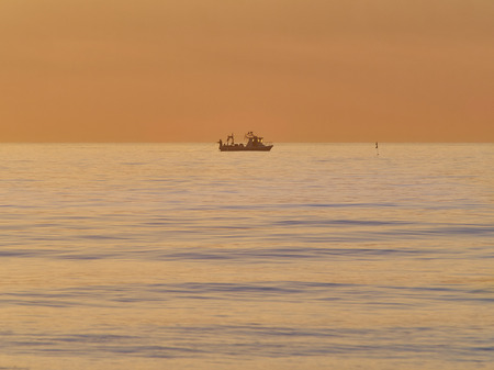fisherman trowing net from a small fishing boat at dusk, calm sea orange skyの写真素材