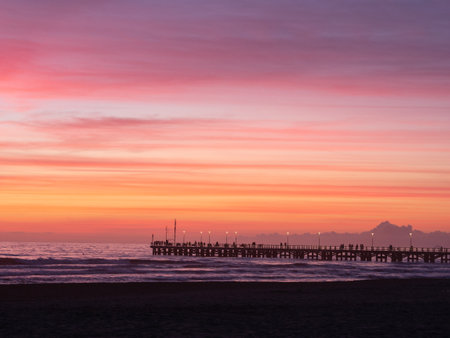Forte dei Marmi's Pier and people walking in a wonderful sunset in the background, the sky and the sea are colored in pink, orange, red, blue and purpleの写真素材