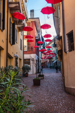 luino, Italy - 12-06-2022: Beautiful street in the historic center with colorful shops and hanging red umbrellas in Luinoのeditorial素材