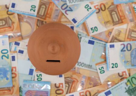 Terracotta money saving box, or piggy bank, on a bed of euro banknotes scattered