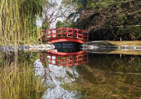 Small wooden red bridge in a woodland with its reflection on a placid river belowの写真素材