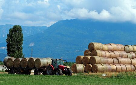 Tractor with hay trailer next to a large stack of round hay bale in a plan under the Alpsの写真素材