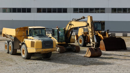 Three yellow construction machineries in a row outside the site. A dump truck, an excavator and a...