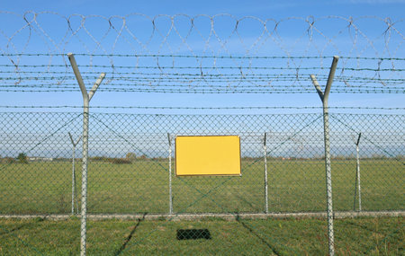 Empty yellow sign outside a double wire mesh barrier with barbed wire on top. Background for copy spaceの写真素材