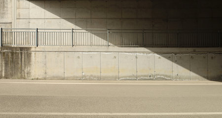Concrete block wall of an underpass with a footpath and a railing in the middle. Side light and shadow. asphalt road in front. No people. Background for copy space.の写真素材