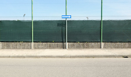 Road directional arrow sign on a concrete sidewalk. Wire mesh fencing with green tarpaulin on behind and asphalt road in front. Background for copy space.の写真素材