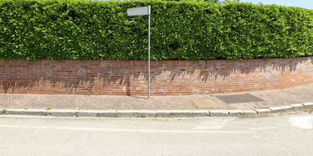Pole with blank street name on a sidewalk. House fencing made of brick wall and hedge on background, asphalt road in front. Background for copy spaceの写真素材