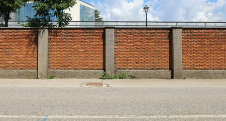Surrounding brick wall with concrete columns with cloudy sky, street lamp and modern building on behind. Urban road in front. Background for copy spaceの写真素材