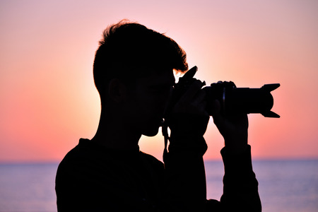 a boy takes pictures on the beach while the sun rises, against the light